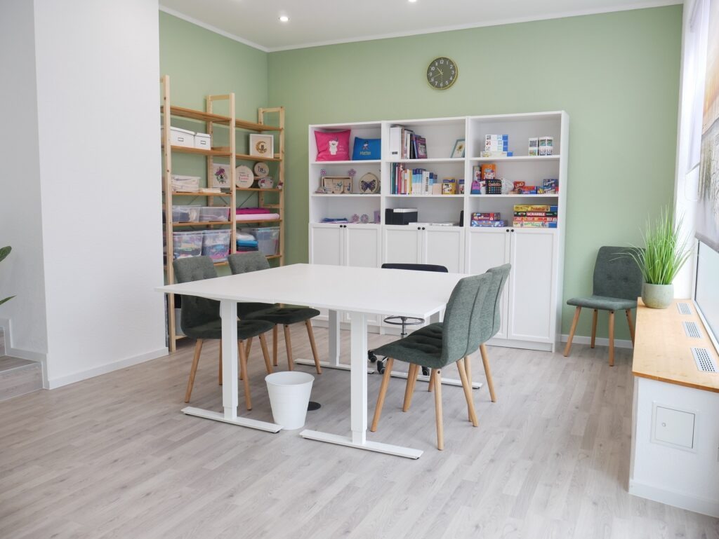 Therapieraum mit Gruppentisch und Materialschrank mit ergotherapeutischen Medien wie z.B. Gesellschaftsspiele, Kreativmaterial und Puzzle.